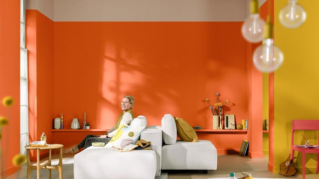 Vīrietis guļ uz modernas pelēkas dīvāna viesistabā ar spilgti oranžām sienām un plauktiem